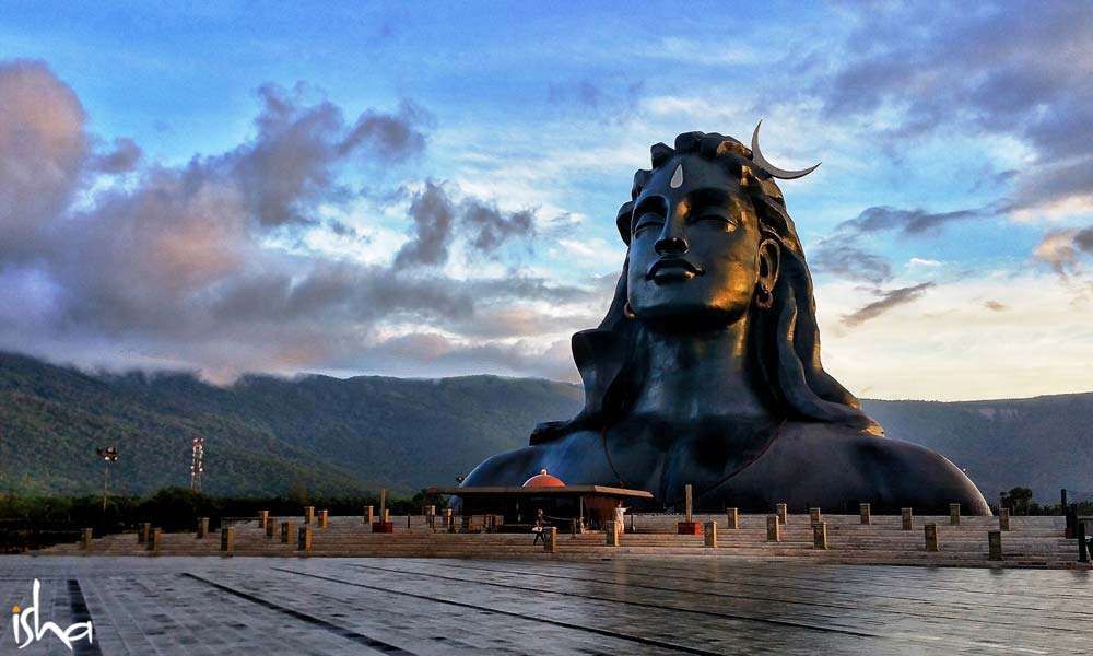 Adiyogi Shiva Statue from an 45 degree angle, with green mountains and cloudy, blue sky in the background.