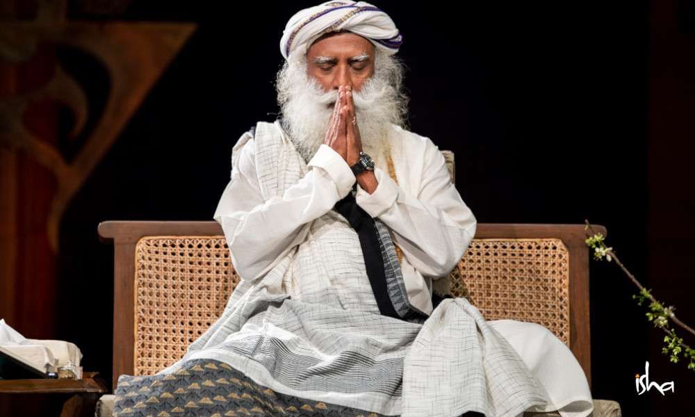 Close up of Sadhguru holding namaskar, eyes close, sitting on chair.